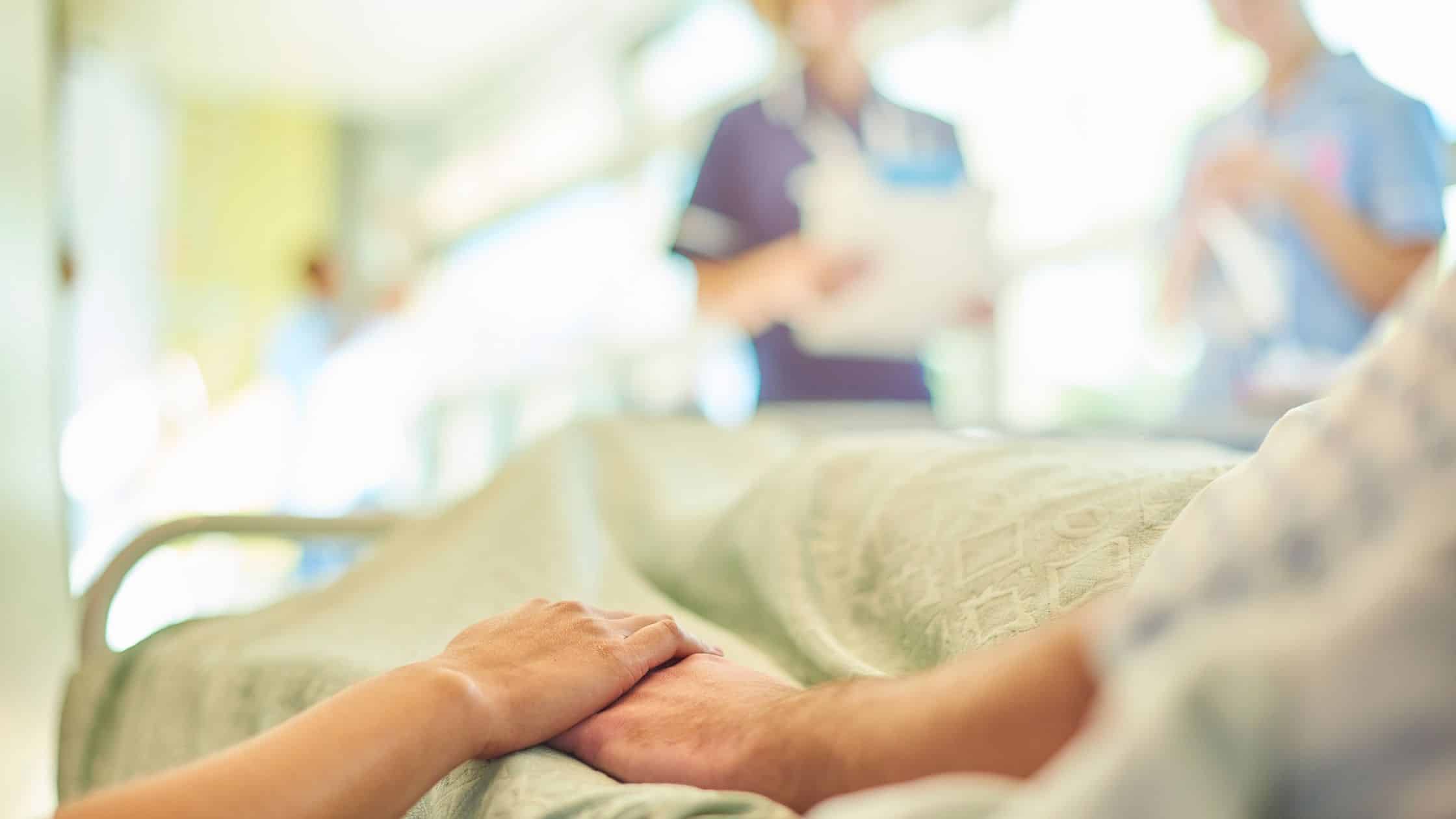 An elderly person in a hospital holding the hand of someone they love.