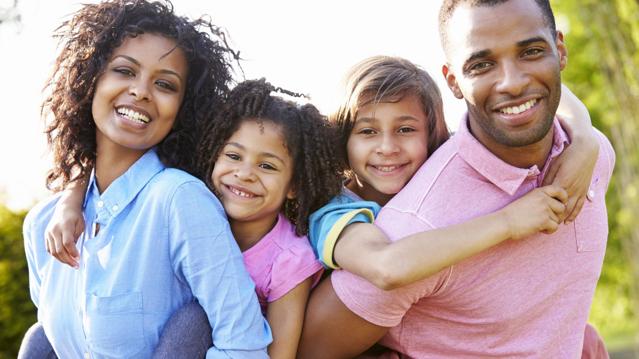 A family smiling as the parents give piggy-back rides to their kids.