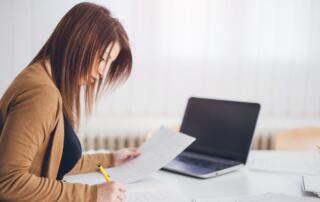 A woman working on paperwork next to her open laptop.