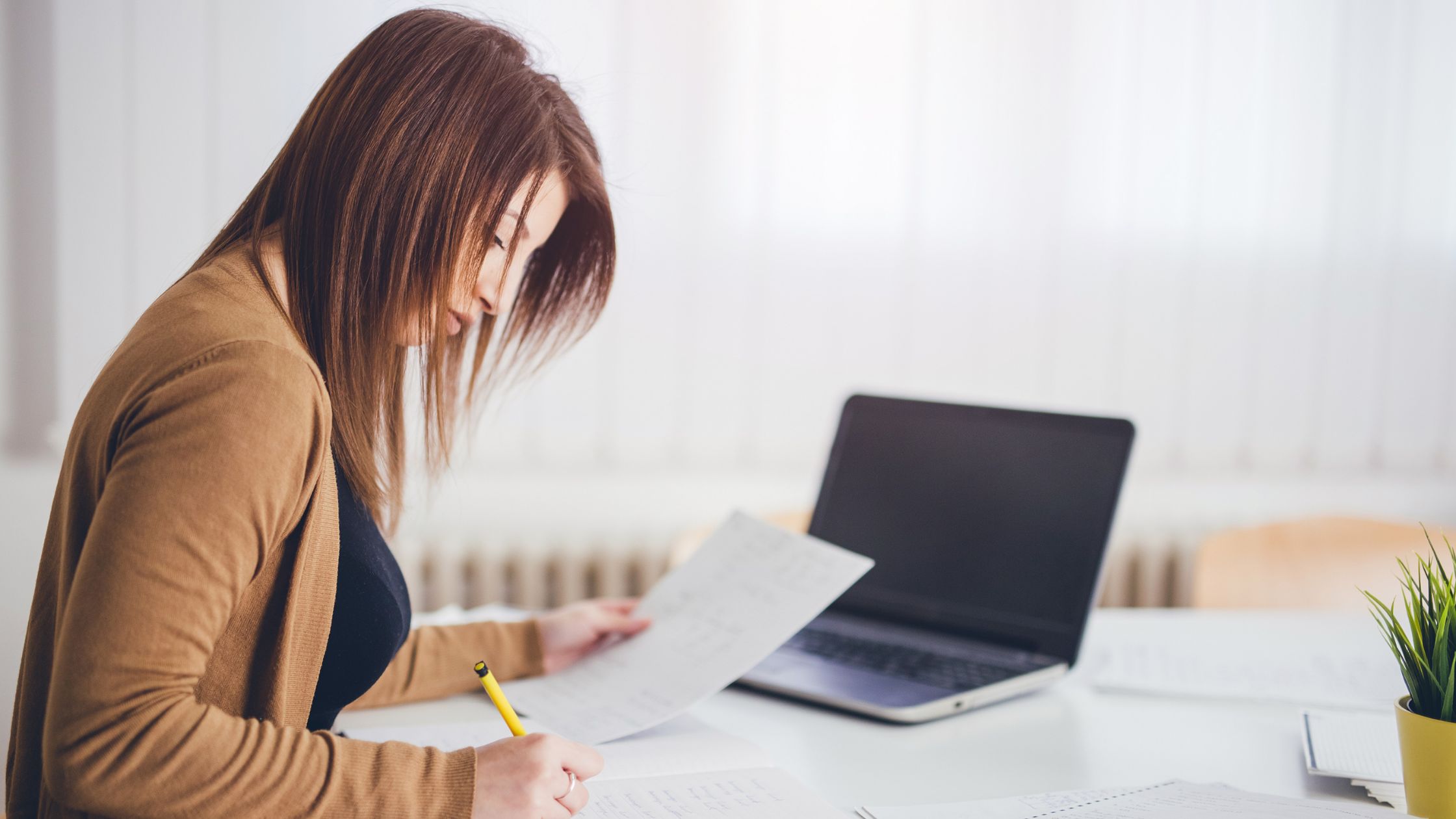 A woman working on paperwork next to her open laptop.