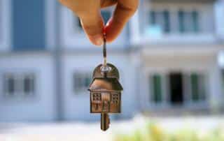 A person holding a set of keys with a house in the background.