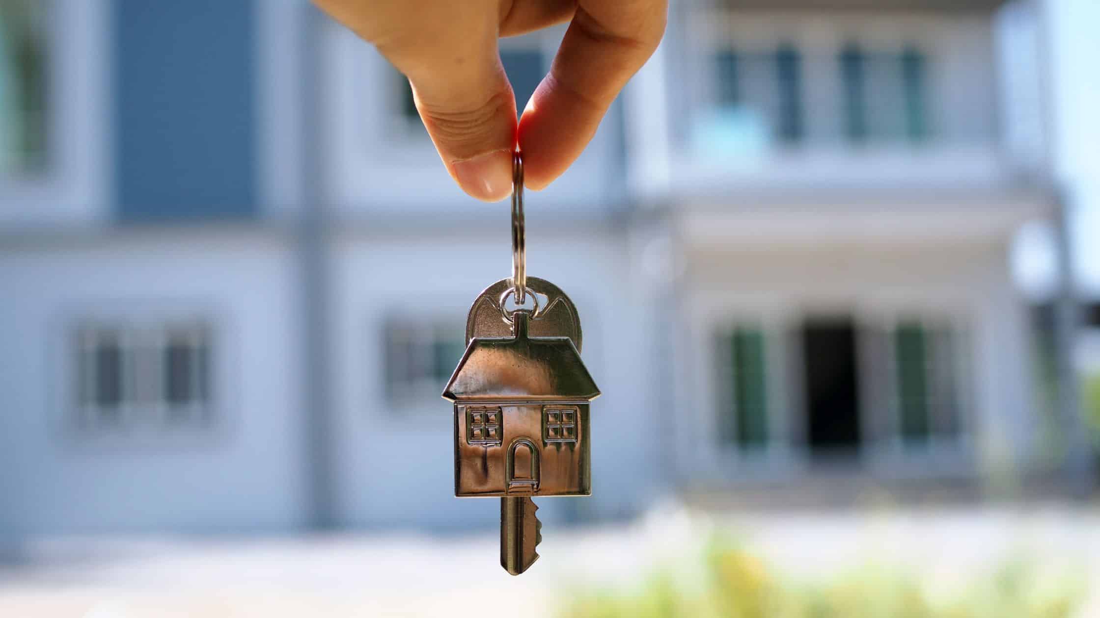 A person holding a set of keys with a house in the background.