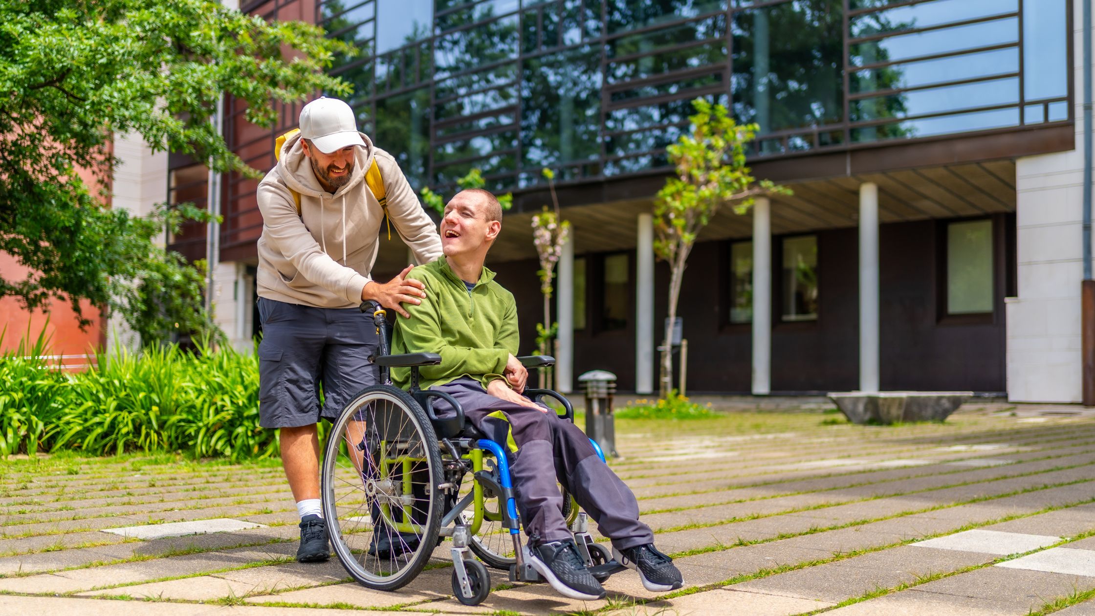 A man pushing a disabled man in a wheelchair, both of them talking and laughing together.