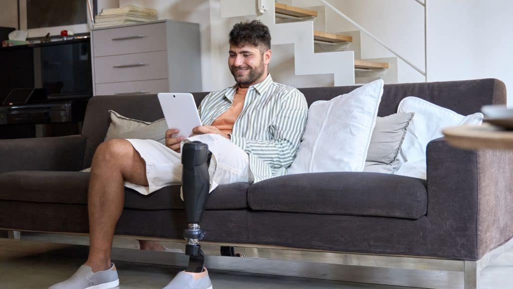 A man with a prosthetic leg sitting on his couch watching something on his tablet.